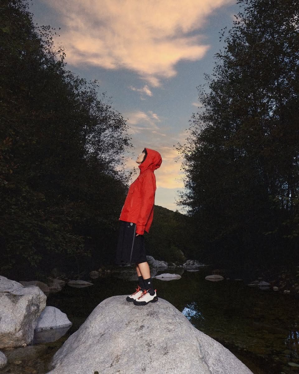 Person standing on a rock near water at dusk wearing outdoor gear