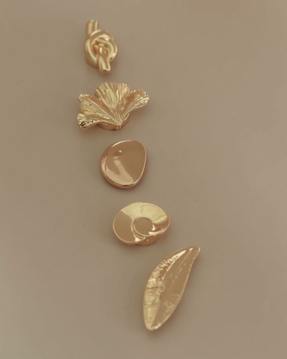 Five gold metallic charms including knot, ginkgo leaf, teardrop stone, spiral shell, and leaf shape