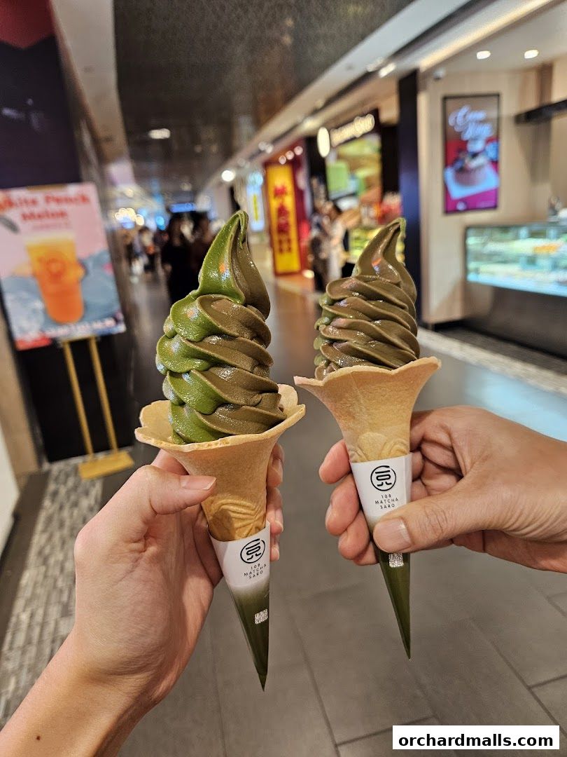 Two hands holding matcha soft-serve ice cream cones in a shopping mall.