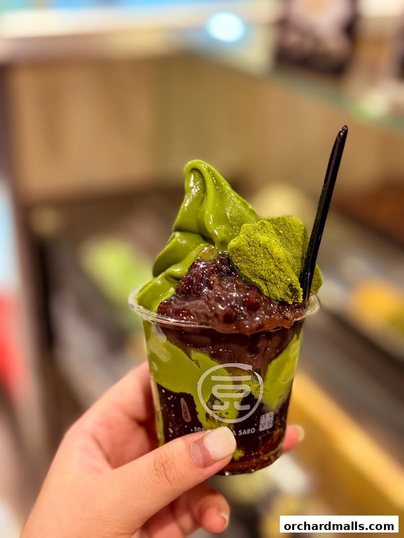 A hand holds a clear cup filled with matcha soft serve, red bean paste, and matcha powder.