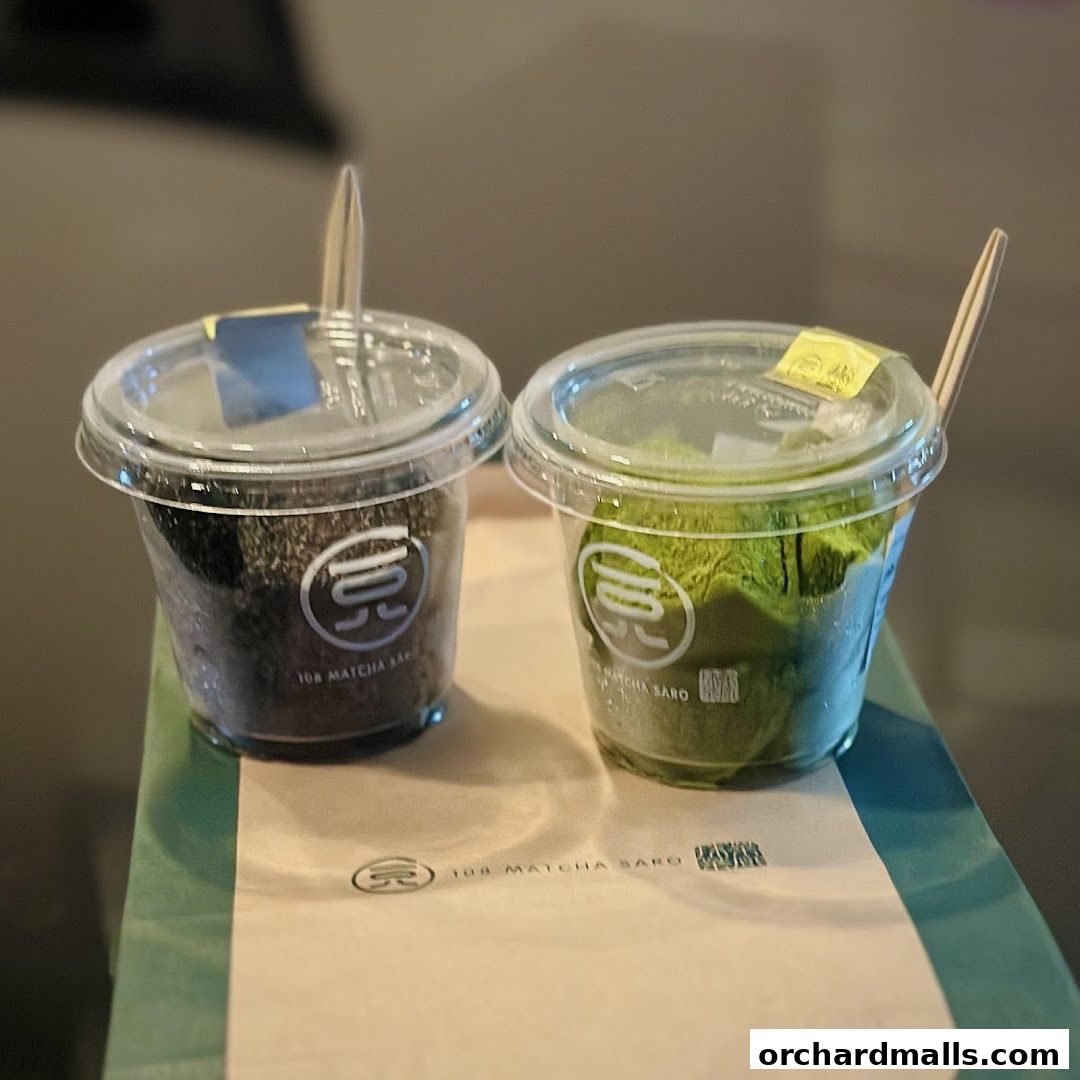 Two clear cups with matcha powder and a dark powder, each with a wooden stirrer.