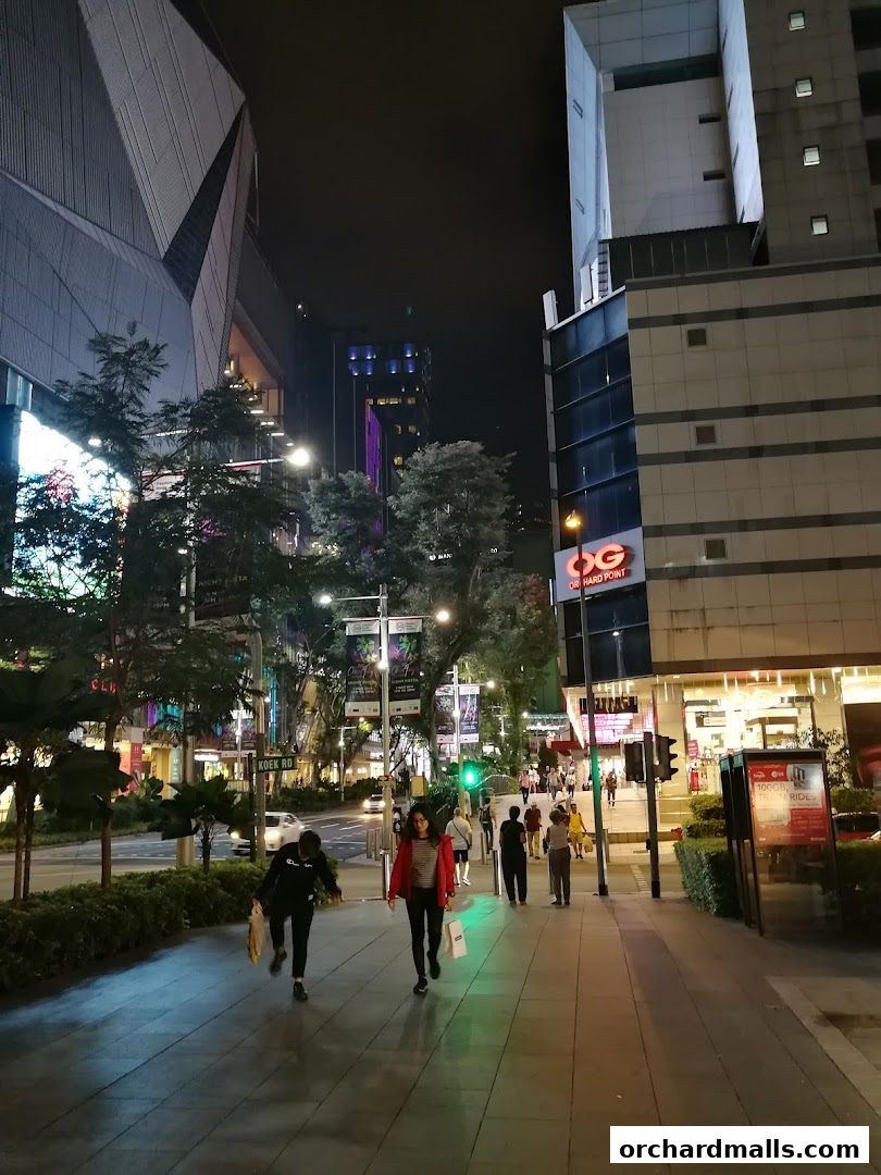 People walking on a sidewalk at night in a city with illuminated buildings and shops.