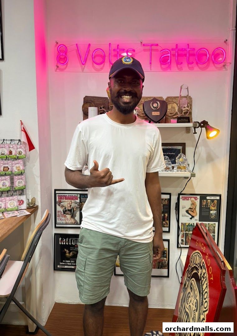 A man stands in front of a neon sign that says '8 Volts Tattoo'.