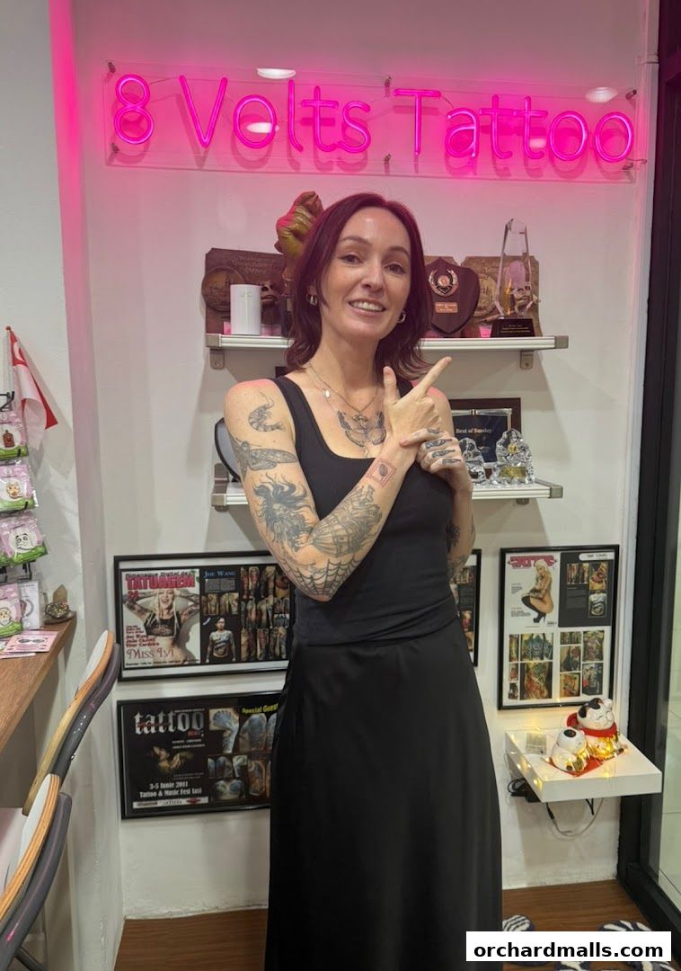 A woman with tattoos stands in front of the '8 Volts Tattoo' sign, pointing.