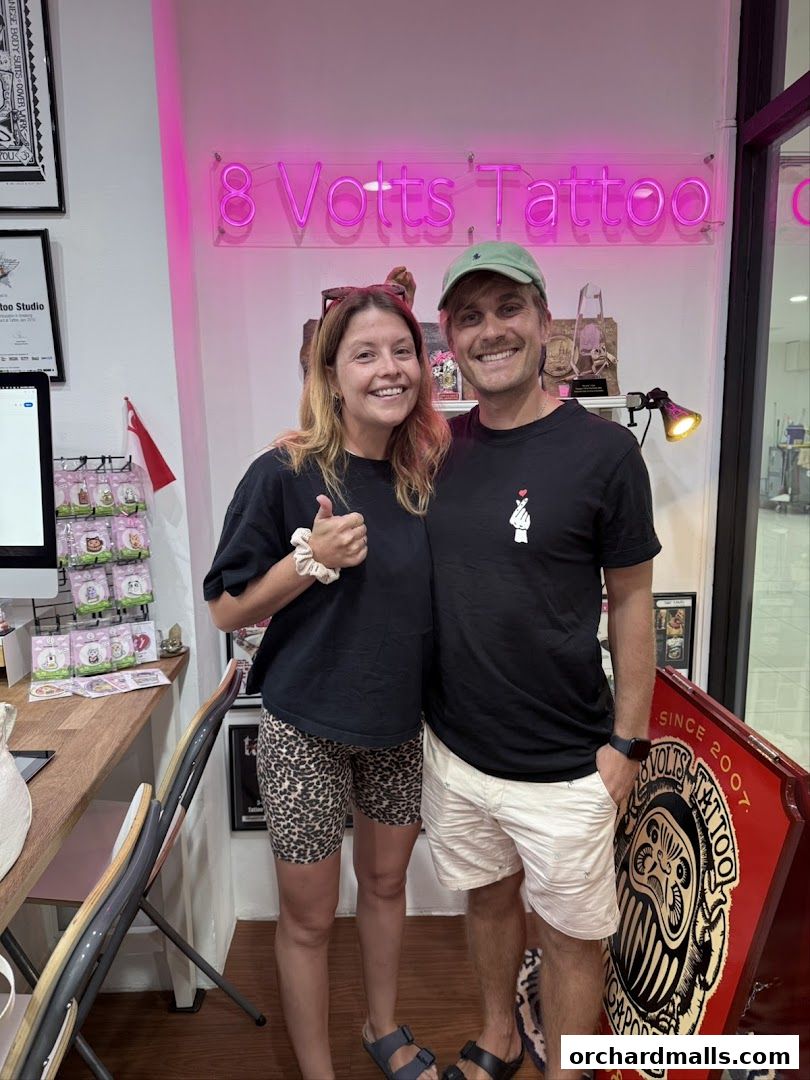 Two smiling people pose in front of a neon sign for '8 Volts Tattoo' in Singapore.