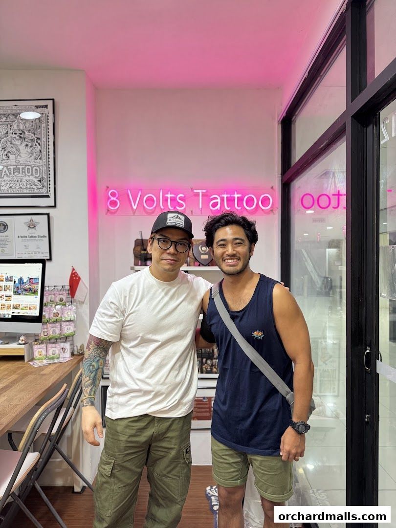 Two men smiling in front of the '8 Volts Tattoo' neon sign.