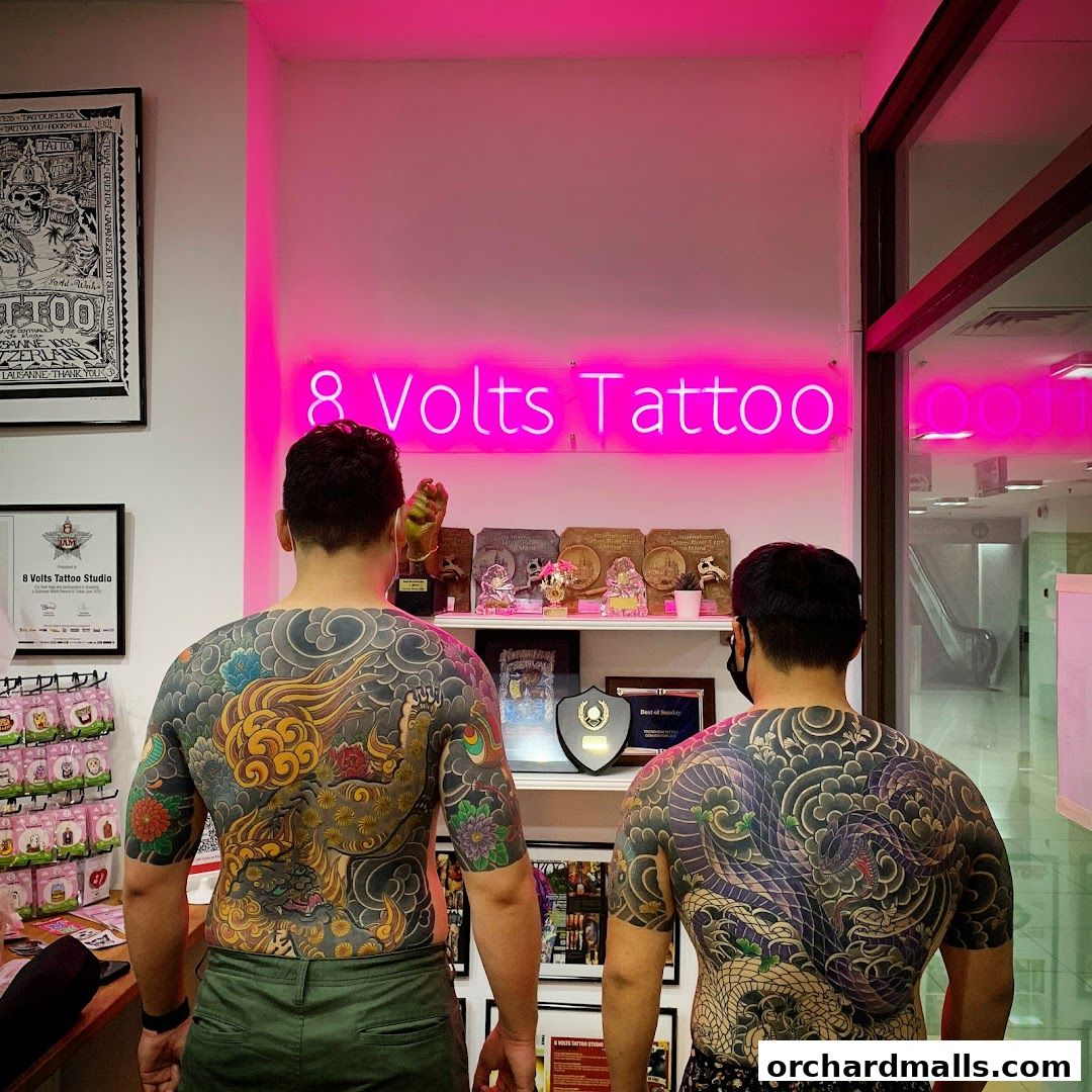 Two individuals display intricate full-back tattoos in a brightly lit tattoo studio.