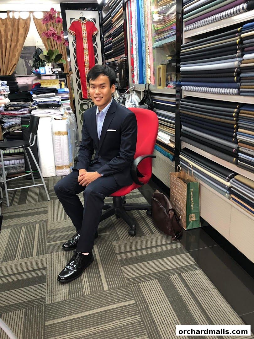 A man in a suit sits in a tailor shop surrounded by fabric rolls and clothing.