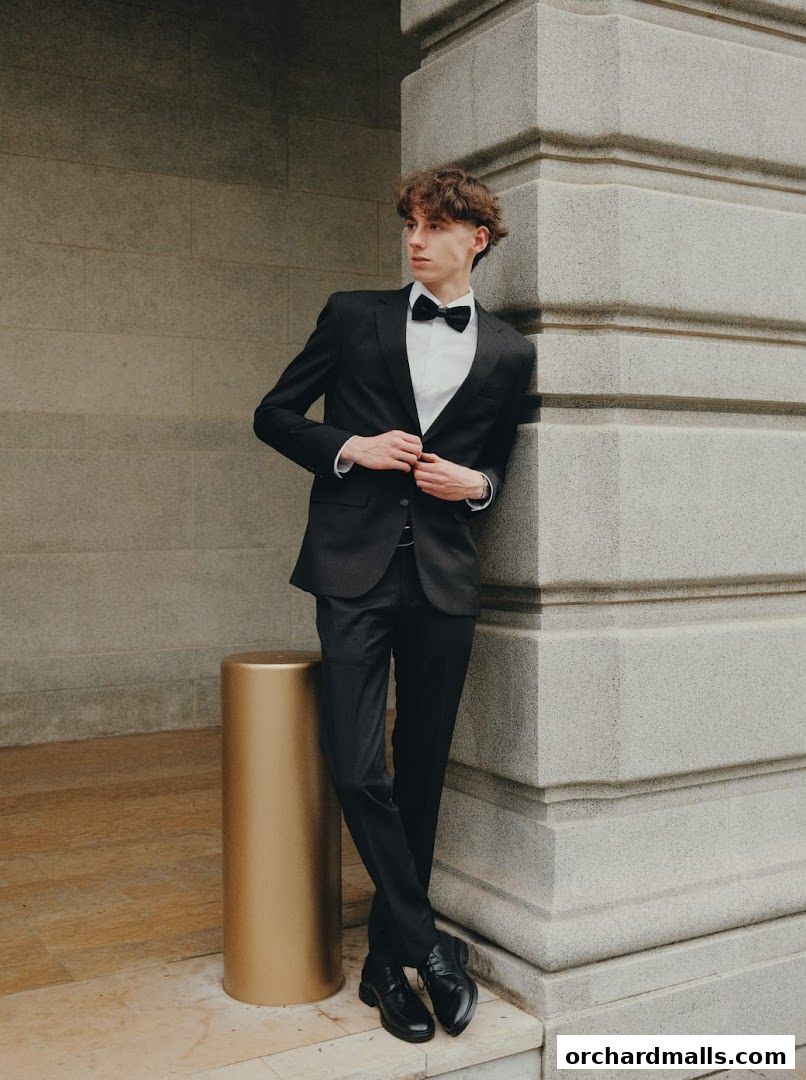 A young man in a black suit and bowtie poses against a stone building.