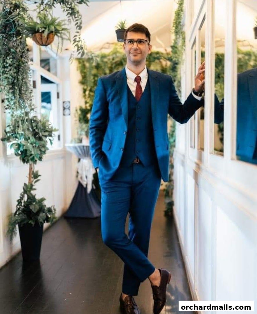 A man in a blue suit stands in a hallway with plants and mirrors.