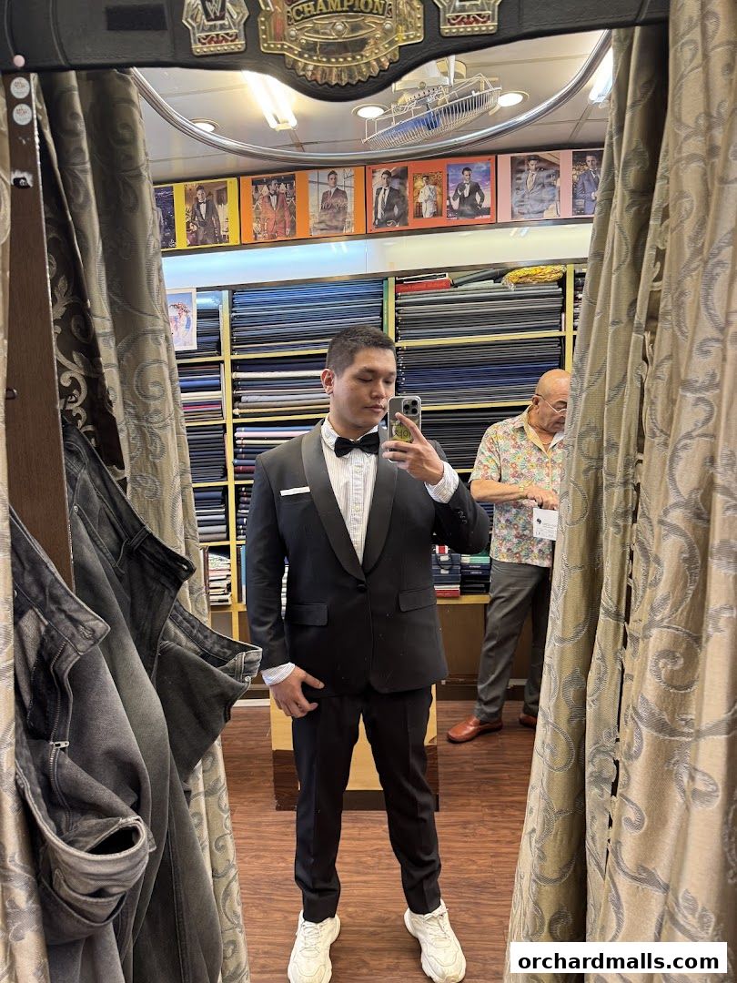 Man in a tuxedo takes a mirror selfie in a tailor shop with fabric rolls in the background.
