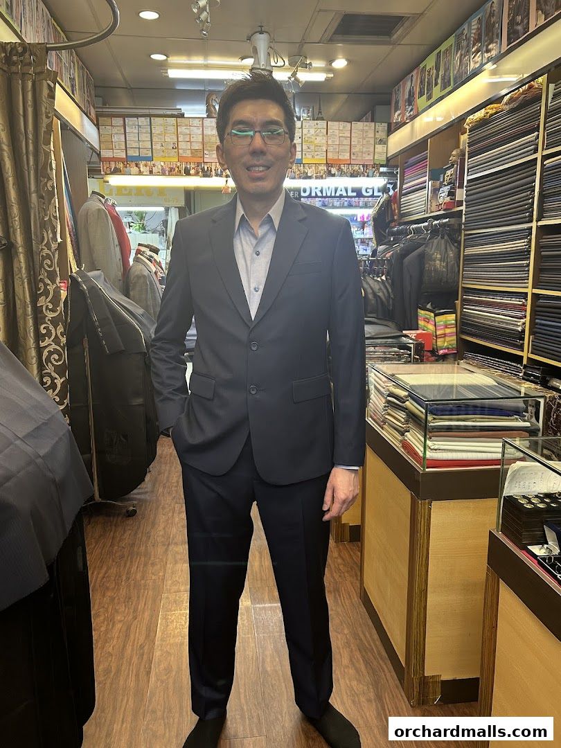 A man in a custom-tailored suit stands inside a tailor shop with fabric rolls.