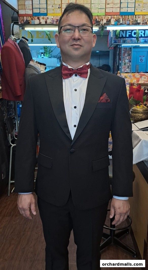 A man in a black tuxedo with a red bow tie stands in a tailor shop.