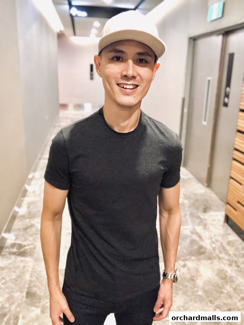 A smiling man wearing a dark t-shirt and a beige cap stands in a modern hallway.