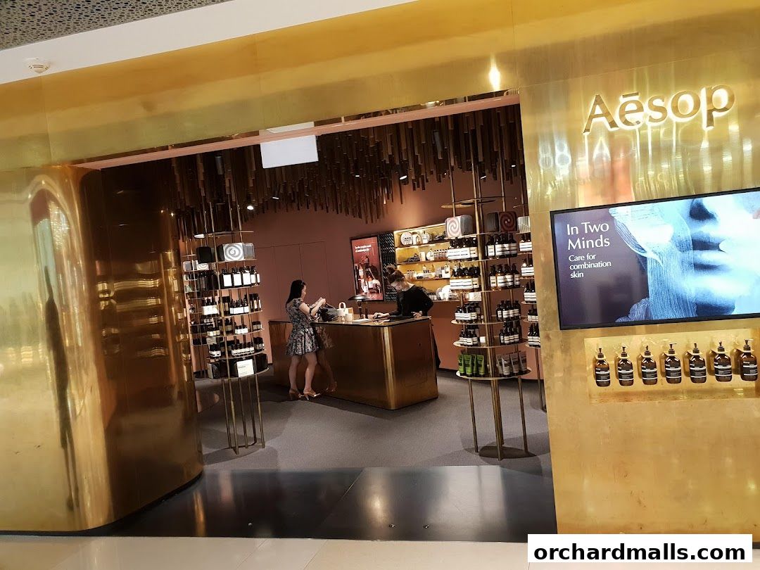 The entrance of an Aesop store with gold accents, displaying products and a promotional screen.