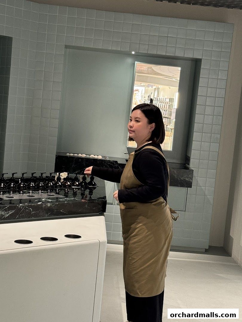 A person in an apron stands near a display of Aesop products.