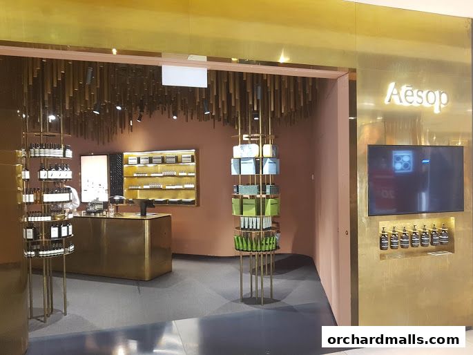 The entrance of an Aesop store with gold accents, product displays, and a digital screen.