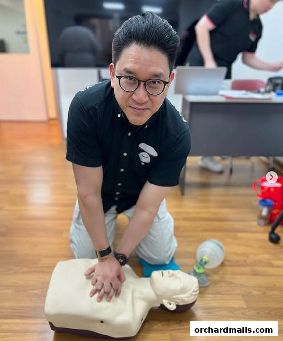 A man in glasses demonstrates CPR on a training dummy in a classroom setting.