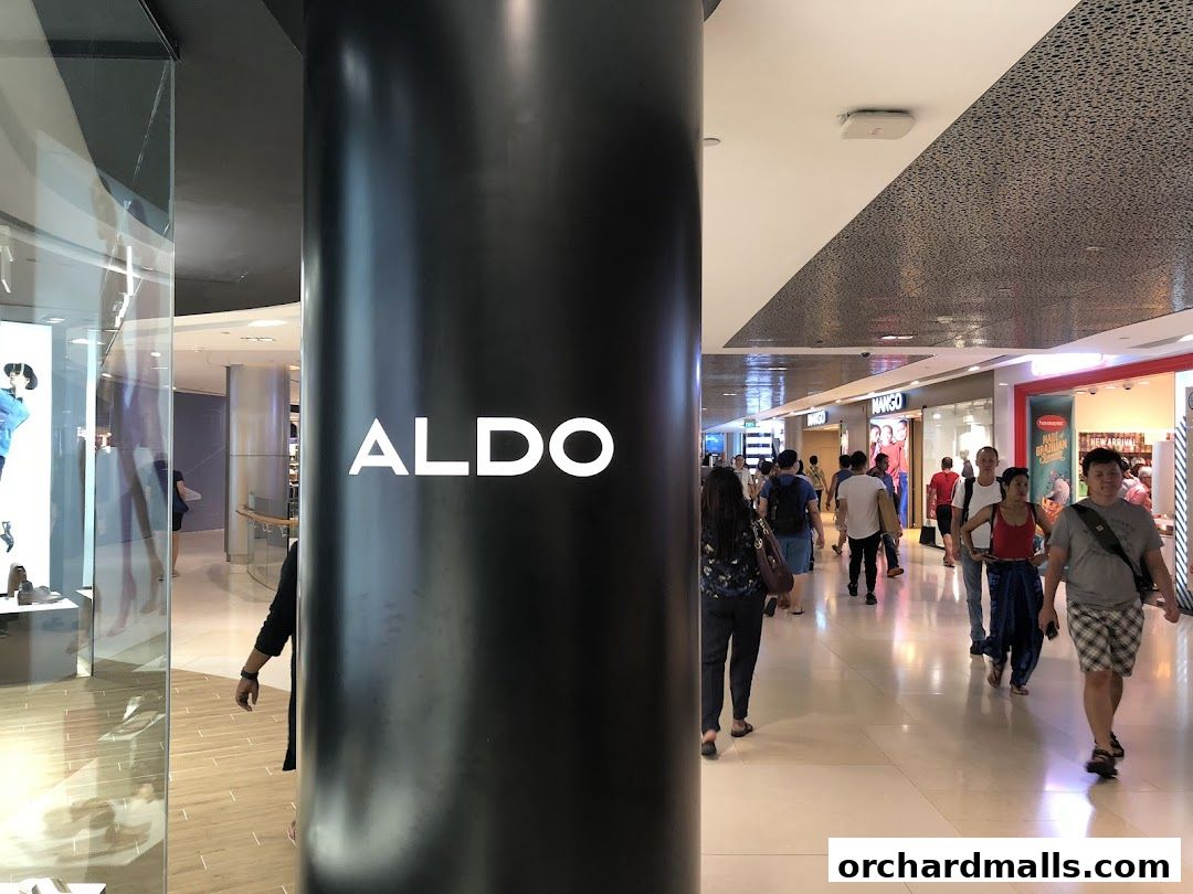 A large black pillar with the ALDO logo is in the foreground, with a busy shopping mall corridor behind it.
