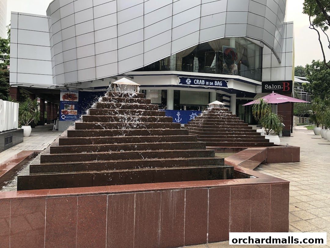 A tiered water fountain in front of a building with shops, including 'Crab in da Bag'.