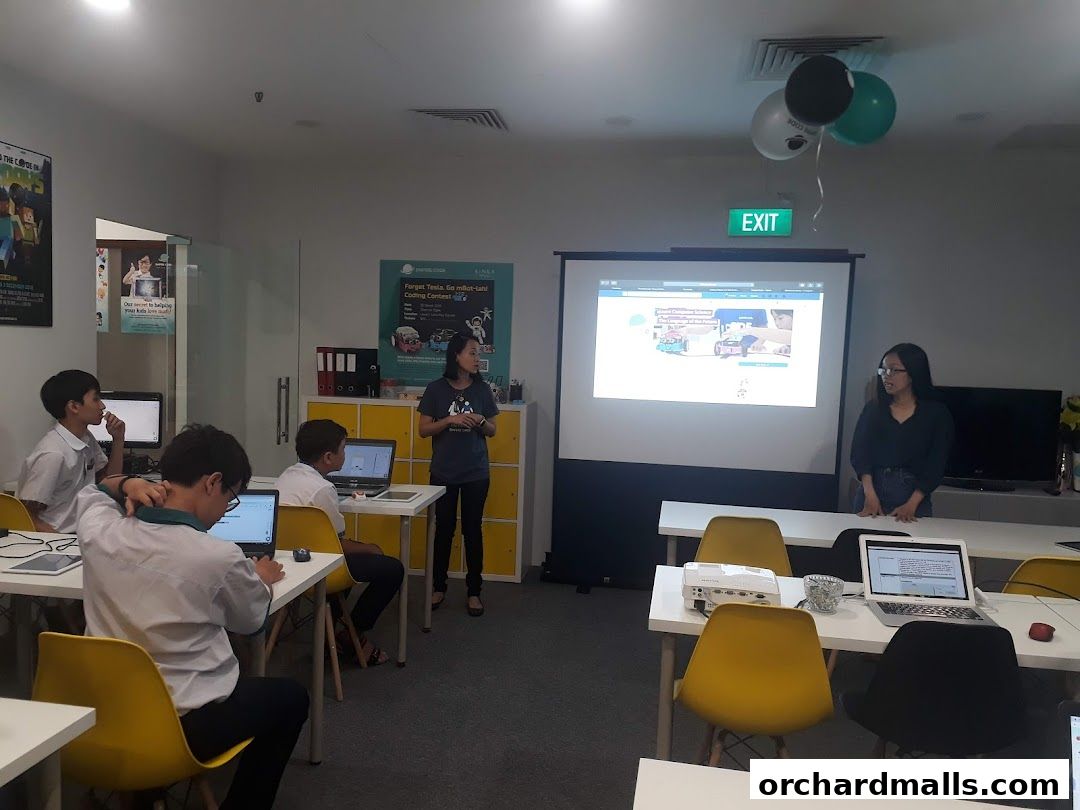Students learning to code in a classroom with a presentation on a projector screen.