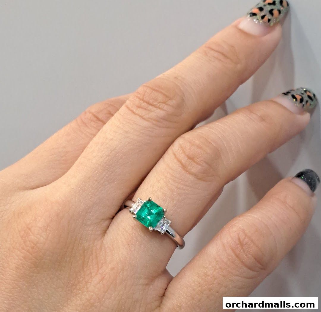 A hand wearing a silver ring with a square emerald and two small diamonds.