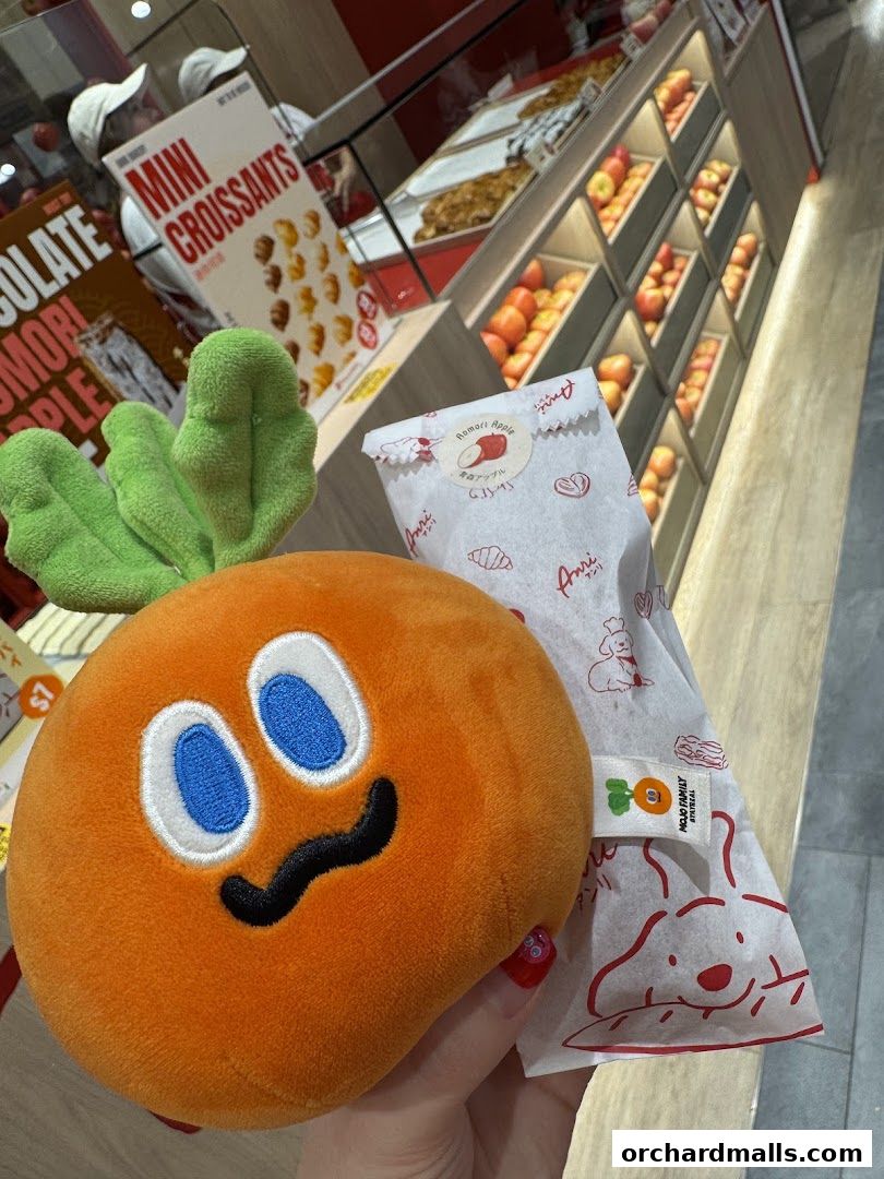 A plush carrot toy held in front of a bakery display with pastries and fruits.