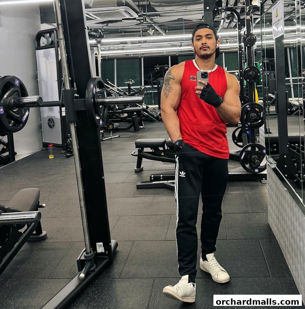 Man in red tank top and black track pants taking a mirror selfie in a gym.