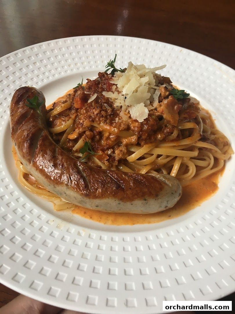 A plate of linguine pasta with bolognese sauce and a grilled sausage, garnished with cheese and herbs.