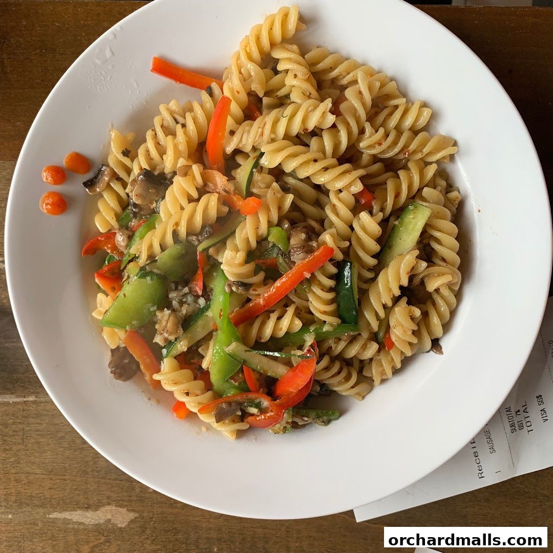 A plate of fusilli pasta with mixed vegetables and mushrooms, served with a side of sauce.