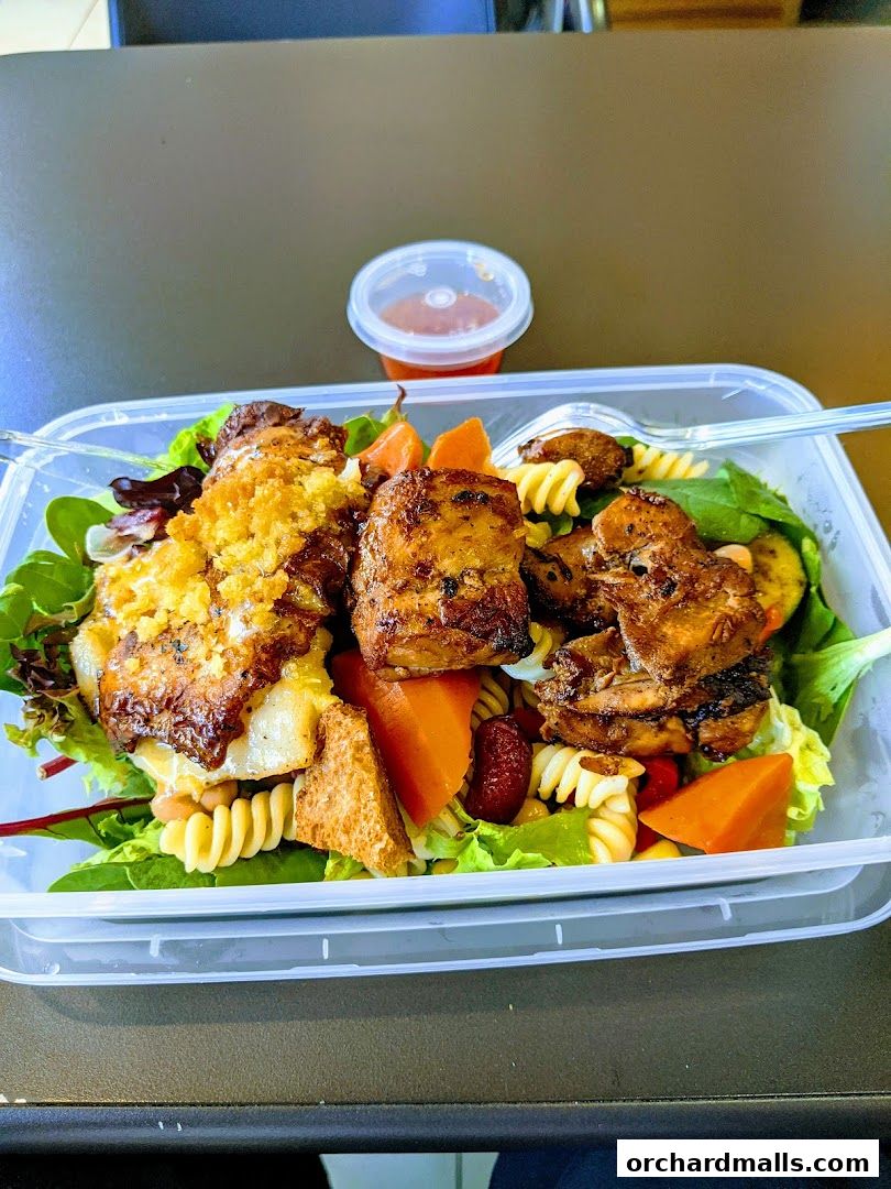 A take-out container filled with a salad featuring grilled chicken, pasta, and vegetables.