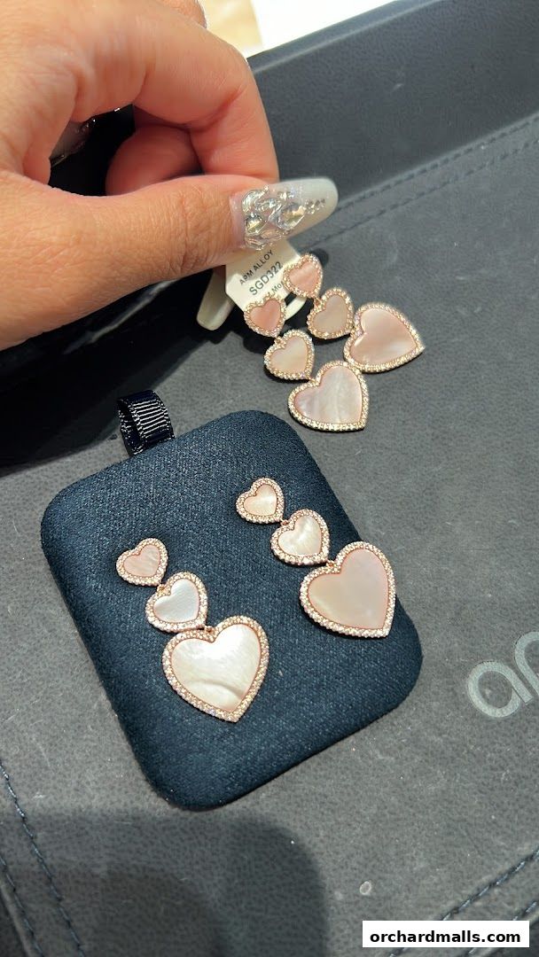 A hand displays delicate heart-shaped earrings with mother-of-pearl and diamond accents.