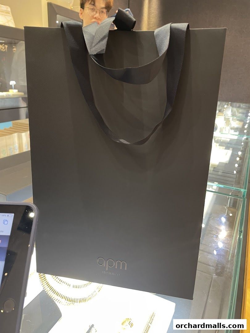 A black APM Monaco shopping bag with ribbon handles sits on a display counter.
