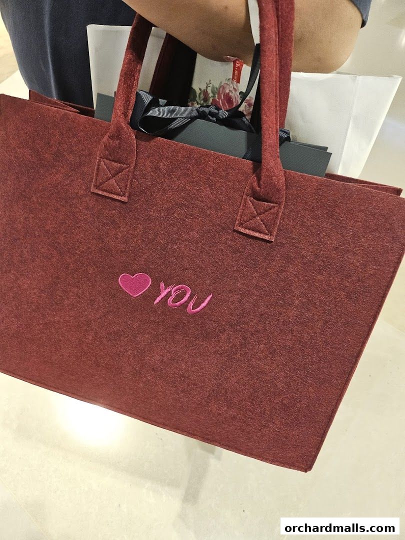 A person holds a maroon felt tote bag with 'I <3 YOU' embroidered on it, filled with shopping bags.
