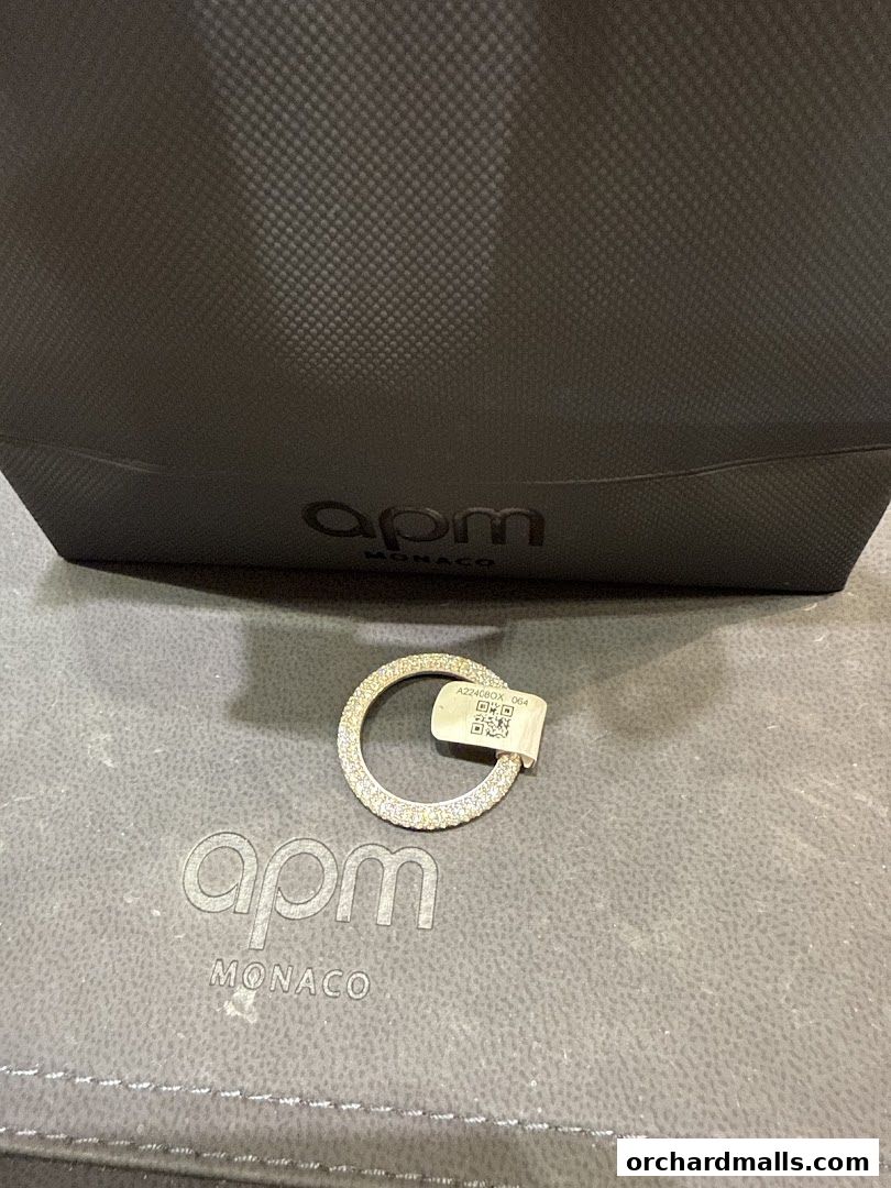 A sparkling ring from APM Monaco is displayed on a branded surface with a shopping bag.