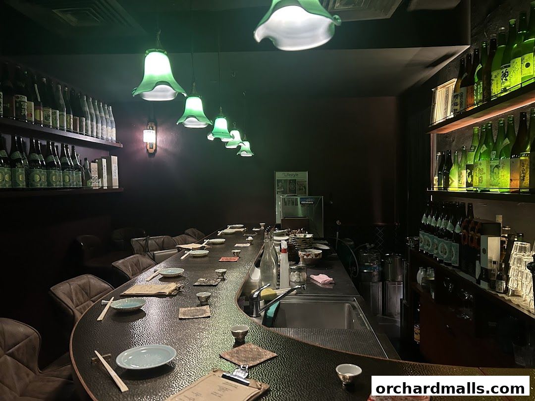 Interior of a dimly lit bar with a long counter, bottles of alcohol on shelves, and green pendant lights.