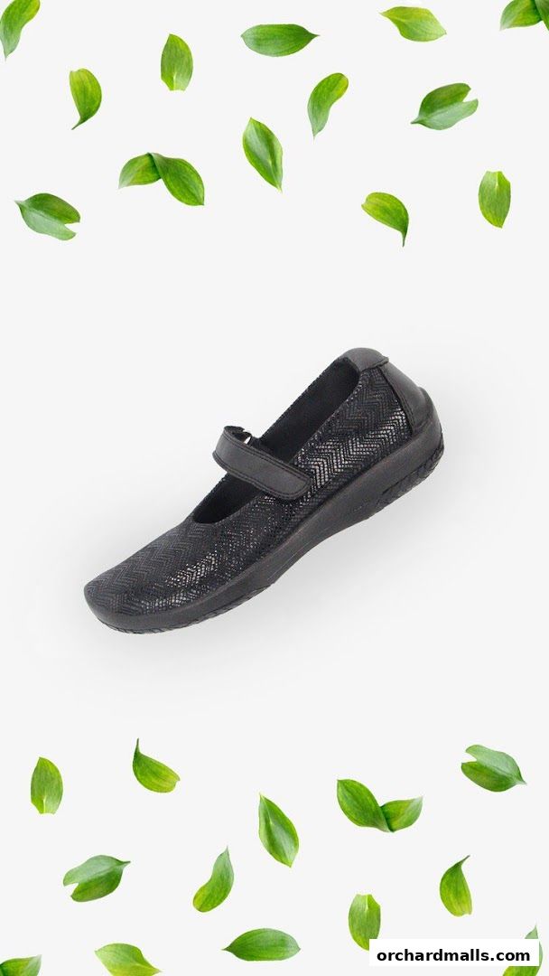 A black Mary Jane shoe with a textured chevron pattern and a velcro strap, surrounded by green leaves.