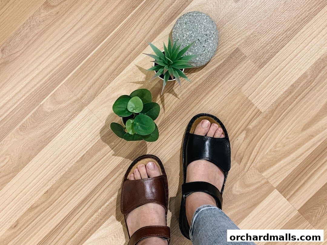 A person wearing brown and black sandals on a wooden floor with plants.