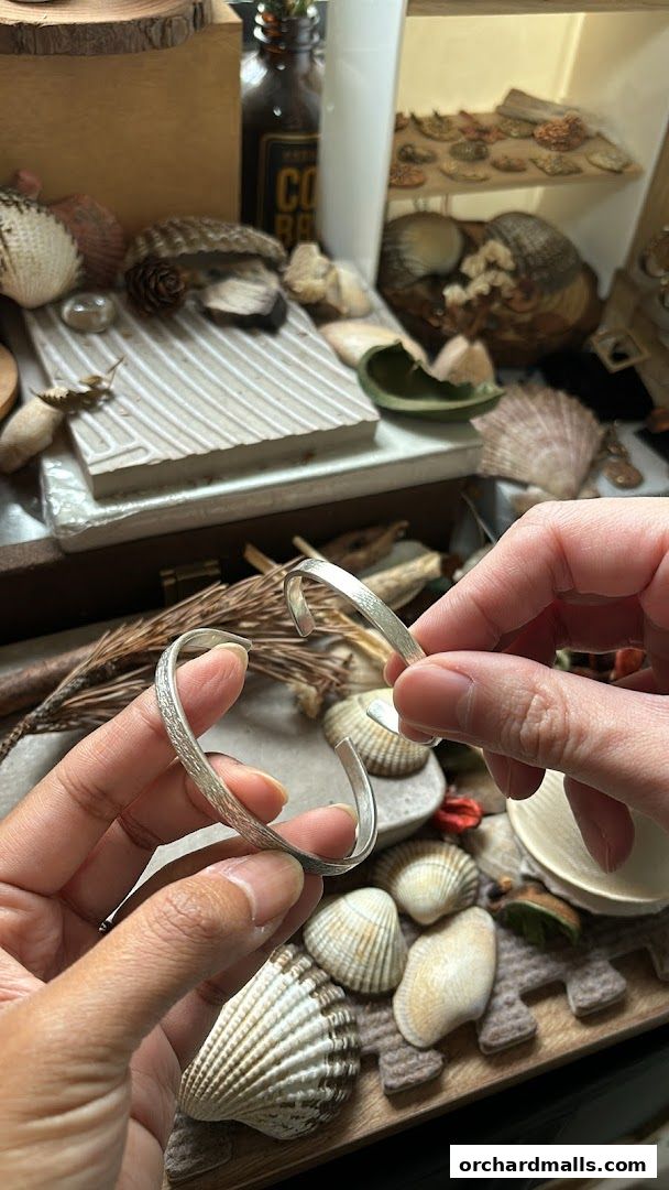 Hands holding a textured silver bracelet amidst shells and natural elements.
