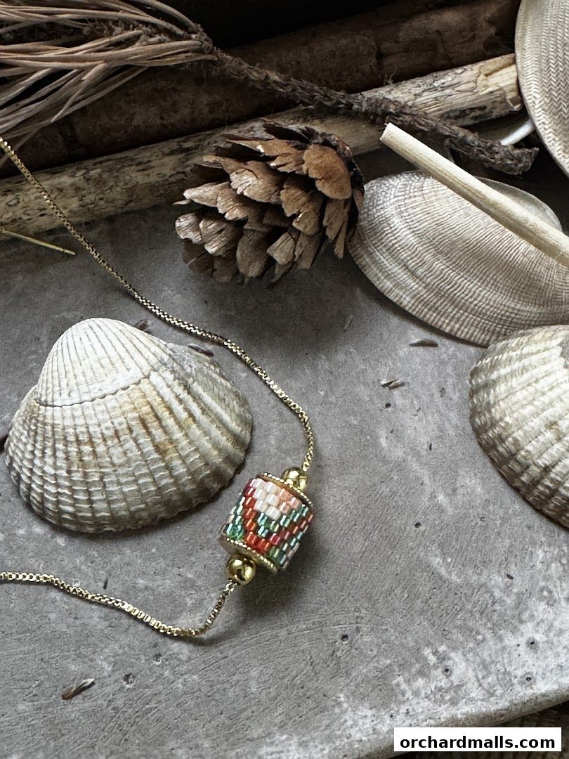 A delicate gold necklace with a colorful beaded pendant rests on a textured surface with shells and pinecones.