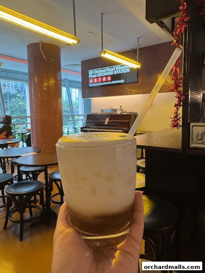 A hand holds a frothy drink in a glass inside ASTRA Deli, with a piano and seating visible.