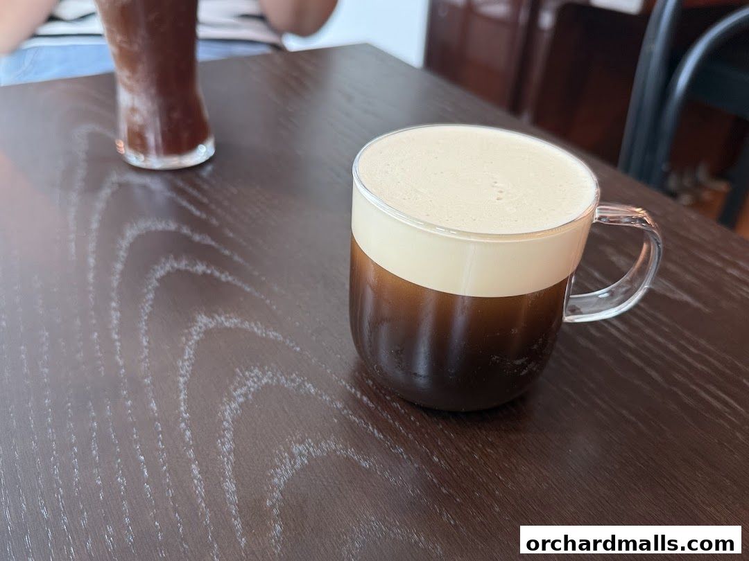 A close-up of a coffee drink with foam on top, served in a clear mug on a wooden table.