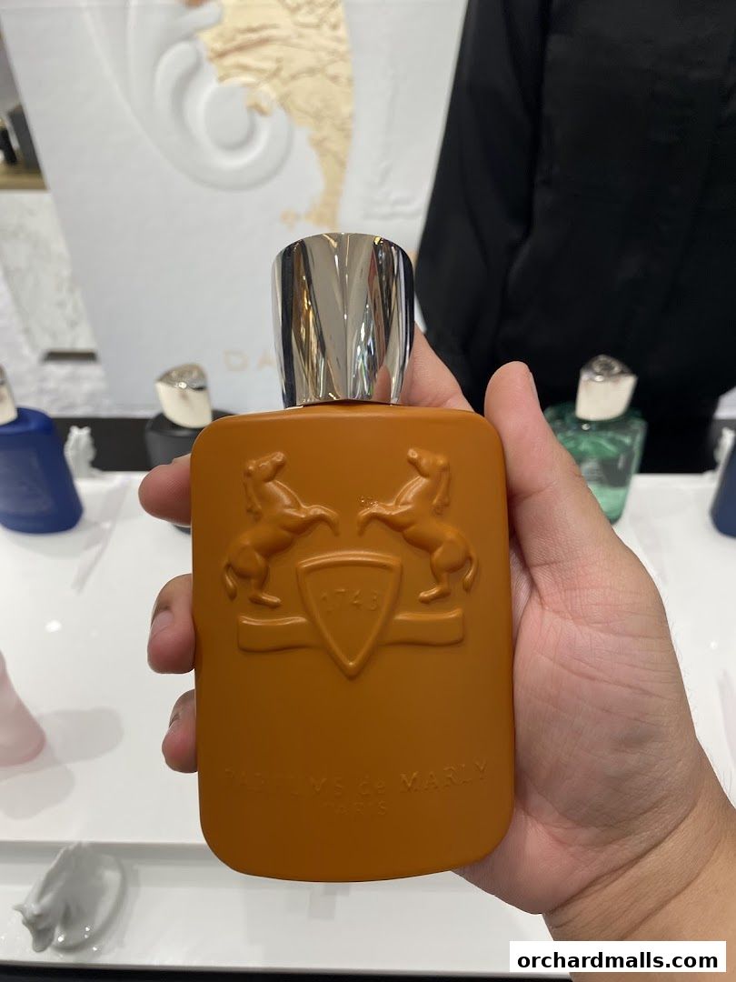 A hand holds an orange perfume bottle with a silver cap, showcasing embossed branding.