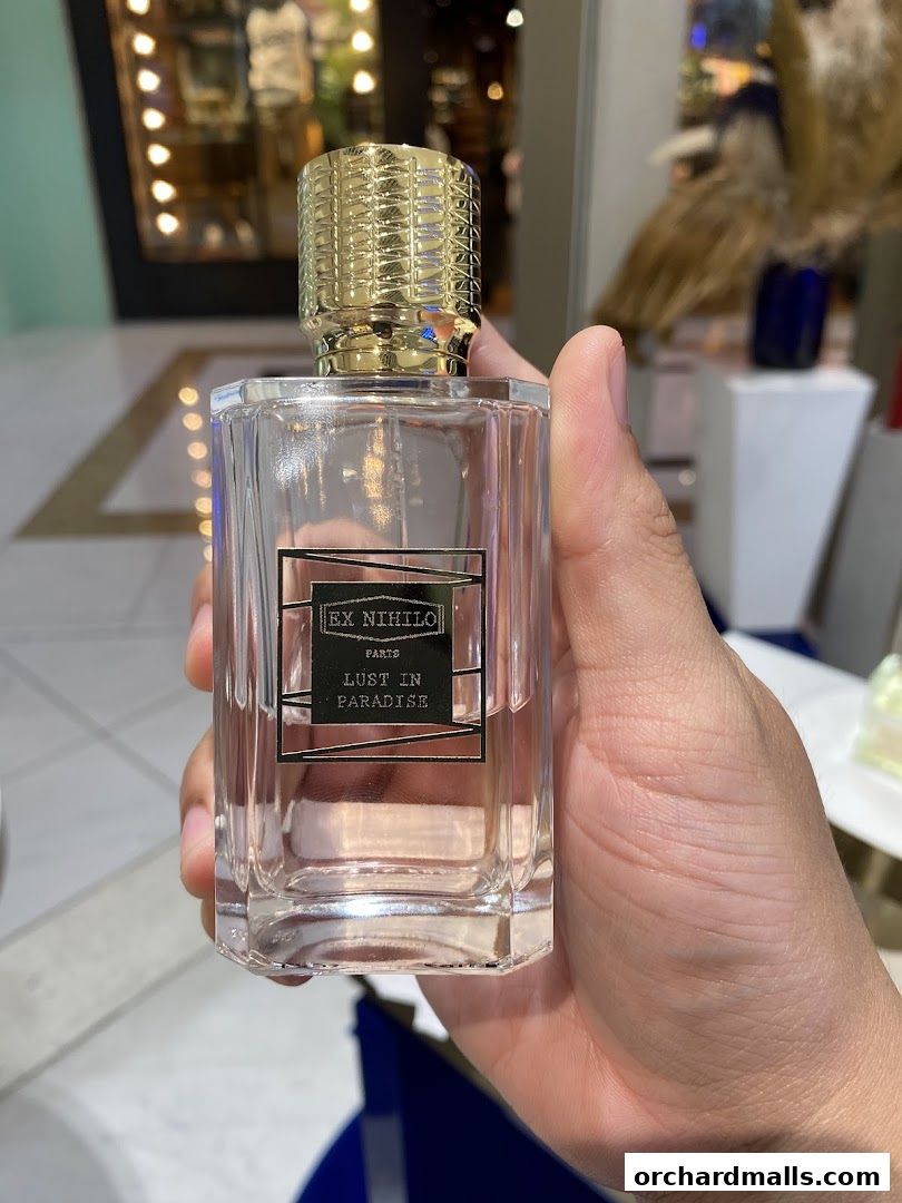 A hand holds a clear glass perfume bottle with a gold cap and black label.