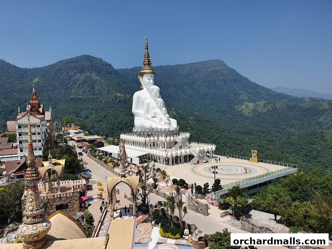 A large white Buddha statue sits atop a hill with lush green mountains in the background.