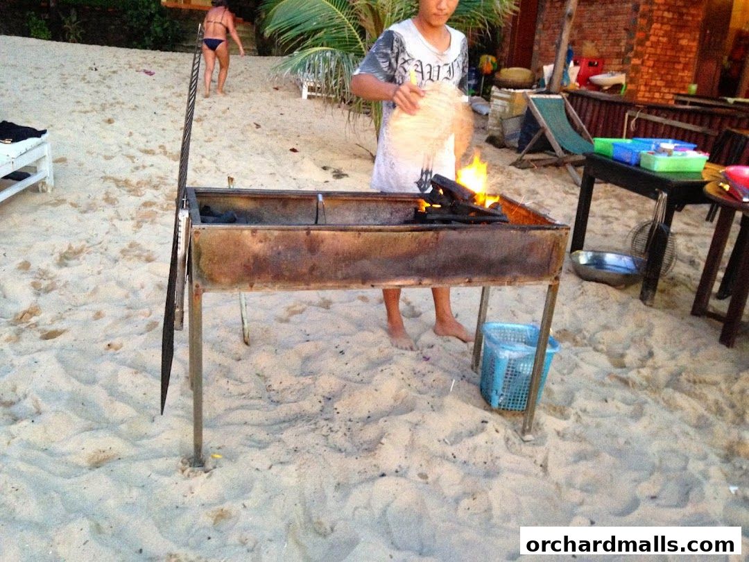 A person is tending to a barbecue grill on a sandy beach with palm trees and seating in the background.