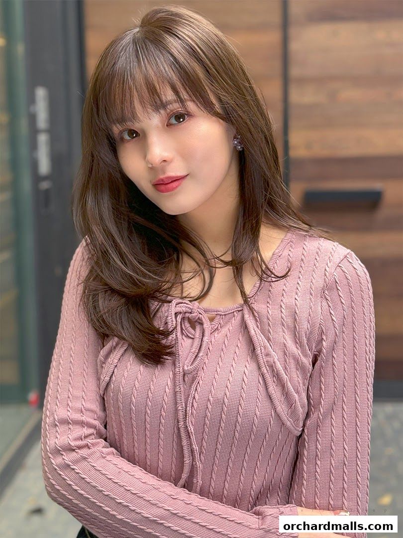 A woman with long, brown hair and bangs poses in a pink knitted top.
