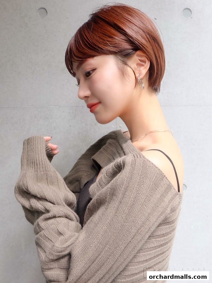 A woman with a stylish short haircut and a warm sweater poses against a concrete wall.