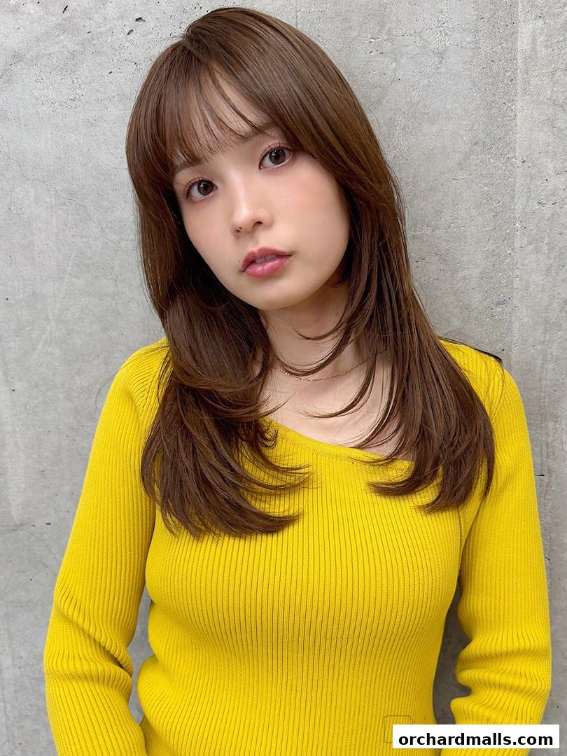 A woman with layered brown hair and bangs, wearing a yellow top, poses against a concrete wall.