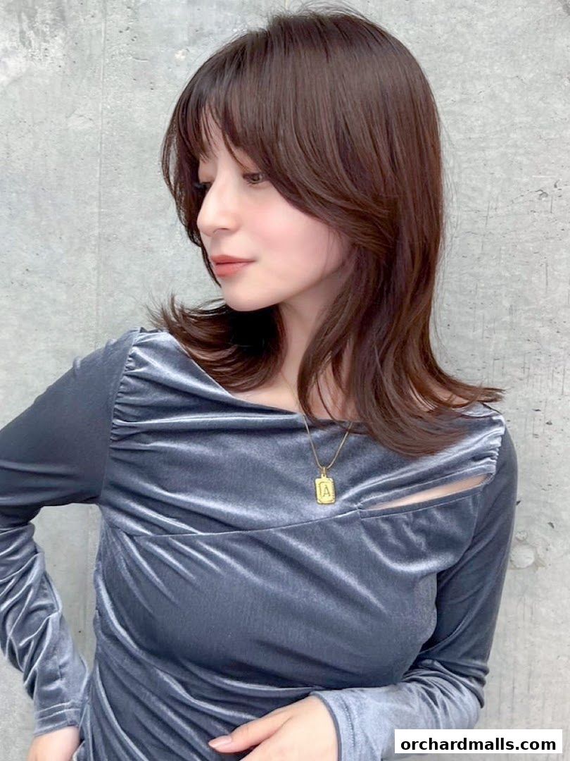 A woman with stylish layered brown hair and a grey velvet top poses against a concrete wall.
